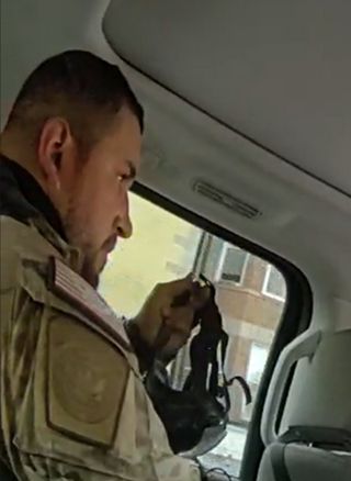 A man sitting in the backset of an SUV. Only his right shoulder and right-profile of his face are visible. In his left hand, he holds a gas mask. On his right shoulder is an American flag patch. He has very short cropped hair and some beard and mustache growth.