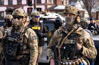 Two male border patrol agents stand in the foreground. Both are masked, dressed in military camo fatigues wearing patches that read "Border Patrol" and "Police BORTAC."

The one on the left wears dark sunglasses and has an identification patch EZ-2, the one on the right EZ-17. 

At least five agents are slightly blurred in the background standing in front of what appears to be a police vehicle.