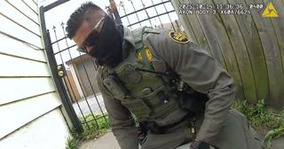 A man in a Border Patrol uniform with a black gaiter mask pulled up to his eyes and wearing sunglasses. He is sitting or kneeling on or near a man he is arresting.