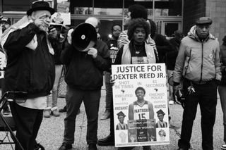 A Black woman with a sign reading "Justice for Dexter Reed Jr. / Shot 96 times in 41 seconds / Executed by Chicago Police / #NoJustice #NoPeace #WeWant #Justice / Black Lives Matter Women of Faith" standing with other protesters and a man with a megaphone outside CPD HQ.