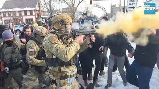 A border patrol agent in military camo fatigues in the foreground fires his launcher directly at the head of a white man a few feet away. The man's face is not visible, obscured by a cloud of orange smoke. Several other neighbors are pictured in the background, mostly obscured by the chemical cloud. Sveum is pictured in the background with American flag patches on his helmet and right arm sleeve.