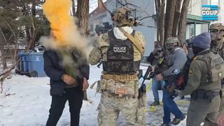 A male Border Patrol agent dressed in military fatigues, a tactical vest, and a ballistic helmet fires a 40mm launcher directly at the head of a Black man. The man's face is not visible, obscured by a cloud of orange gas. The ground is covered in snow, other agents are in view to the right.