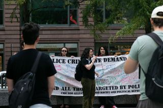 In the foreground, the backs of two protesters. In the background, a speaker with a mic stands in front of activists holding up a white banner with names in colored marker across it. Some banner text is visible, one can read the words “and all those targeted by police.”