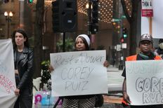 Three protesters in front of city buildings. A Black woman in the middle with a white cap is holding a white sign with “Rest in Power COREY RUIZ” scrawled on it in black marker.