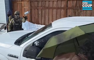 A border patrol agent in military camo fatigues, a mask, and dark sunglasses in the background tosses a canister of tear gas toward the camera, over the top of a white Ford F150 between the agent and camera. Behind him in the background is an agent not facing the camera in a knit cap and black mask.