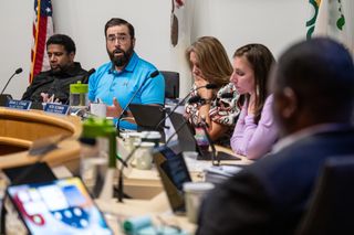 Five trustees sitting at round desk at village meeting. A man in a blue polo is speaking and gesturing with his hands