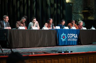 Seven people sit behind a long table on stage, with a blue CCPSA banner over the center table.