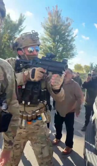 Edgar Vazquez stands directly in front of the camera aiming his launcher off to camera-right. He wears military camo fatigues, mirrored sunglasses, a mask, a ballistic helmet, a tactical vest and a belt holding a colorful array of at least a half-dozen chemical and foam munitions for the launcher.