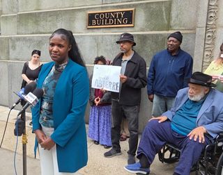 A young Black woman in white pants and long blue coat stands at media microphones with several supporters behind her.
