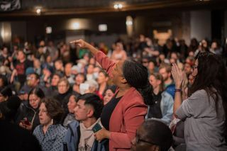 A woman stands amid a crowd and points at someone out of frame, while another woman behind her claps.