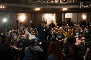 Chicago Alderman Andre Vasquez stands up amid a crowd in a packed venue space, pointing a finger toward CCPSA commissioners (not shown).