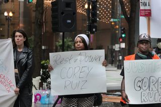 Three protesters in front of city buildings. A Black woman in the middle with a white cap is holding a white sign with “Rest in Power COREY RUIZ” scrawled on it in black marker.