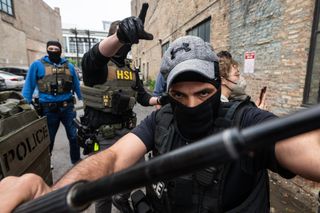 close up of masked ICE agent in an alley pushing back against viewer/photographer with a baton