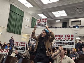 One individual in the foreground stands holding up a sign and appears to be shouting, flanked by other people holding the same sign.