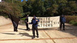 three men in matching black pants and blue jackets holding banner that reads white history matters. james is holding a black flag with the group's logo on it